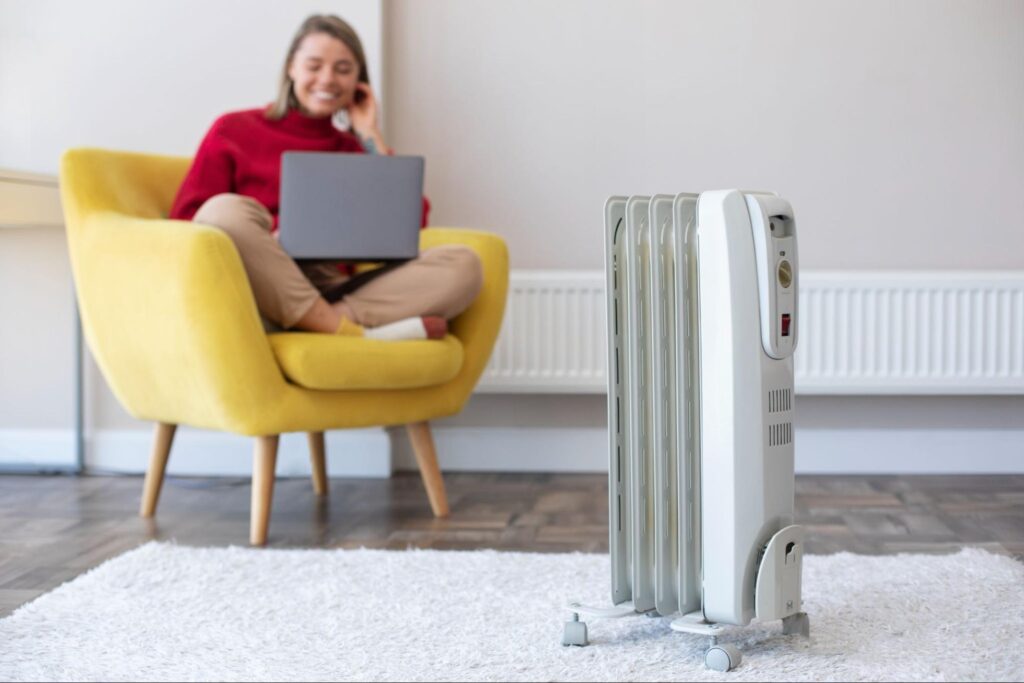 Woman sitting on a yellow chair with a laptop while a portable electric heater runs on a rug in a bright living room.